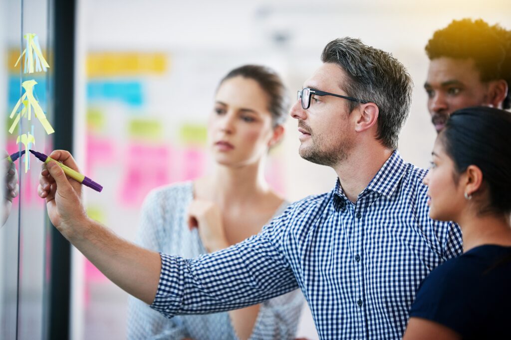 four employees in a meeting with a man writing notes on a post-it adhered to a glass wall.